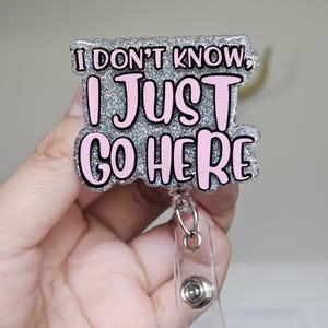 Könnte beinhalten: Ein glitzernder Ausweis-Rollenhalter mit dem Text "I DON'T KNOW, I JUST GO HERE" in rosa und schwarzer Schrift. Der Ausweis-Rollenhalter hat einen transparenten Kunststoffclip und ein einziehbares Kabel.