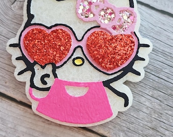 May include: A white cartoon cat-like character with black outlines, wearing heart-shaped red glitter sunglasses and a pink dress. A pink bow with glitter accents sits atop the head. The character is on a light wood surface.