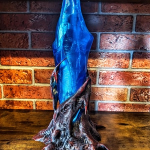 Peut inclure: Une grande lampe en forme de cristal bleu translucide éclairée de l'intérieur. La base est fabriquée dans un matériau sombre, texturé et organique avec des reflets brun rougeâtre qui semble envelopper le bas de la structure en cristal bleu.