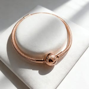 May include: Rose gold bracelet with a round clasp. The bracelet has a textured, snake-like chain design. The clasp features an engraved pattern. The bracelet is displayed on a white surface.
