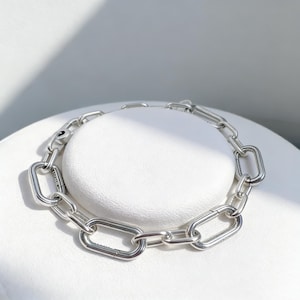 May include: A silver chain bracelet with large, rectangular links. The bracelet is made of a shiny, metallic material and is displayed on a white surface. The links are connected with smaller, rounded links.