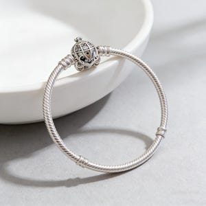 May include: A silver bracelet with a decorative charm. The charm has a detailed, ornate design. The bracelet band has a textured, ridged pattern. The bracelet is displayed on a white surface against a gray background.