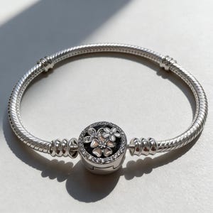 Puede incluir: Una pulsera plateada con un diseño de cadena de serpiente. La pulsera presenta un dije redondo con acentos florales blancos y rosas, rodeado de cristales transparentes. El dije está sobre un fondo negro.