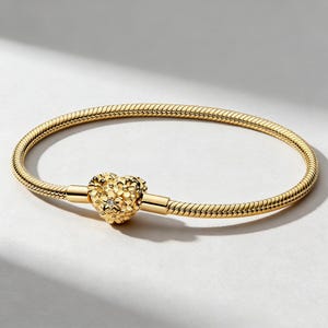May include: A gold bracelet with a snake chain design. The bracelet has a heart-shaped clasp with floral detailing and a small, sparkling accent. The jewellery is displayed on a white surface.
