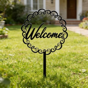 May include: A black metal garden stake with the word "Welcome" in a cursive font. The word is inside a decorative circular frame with scrollwork. The stake is in a grassy yard.