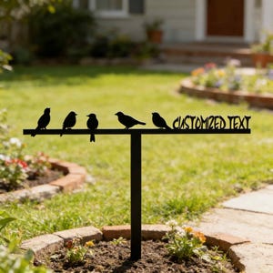 May include: Black metal garden stake featuring five bird silhouettes perched on a horizontal bar. The words "CUSTOMIZED TEXT" are cut out on the right side of the bar. The stake is in a garden setting with flowers and grass.
