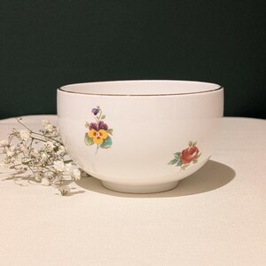 Könnte beinhalten: Eine weiße Keramikschale mit goldenem Rand und floralen Designs. Die Schale zeigt ein Stiefmütterchen und eine Rose, beide in leuchtenden Farben. Die Schale steht auf einer hellen Oberfläche, mit einem Zweig weißer Blumen links.