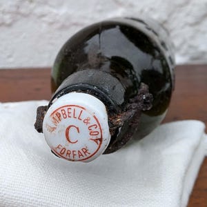 May include: A vintage dark green glass bottle with a white cap bearing the text "Campbell & Co FORFAR" in red. The bottle has a metal clasp and rests on a white cloth.