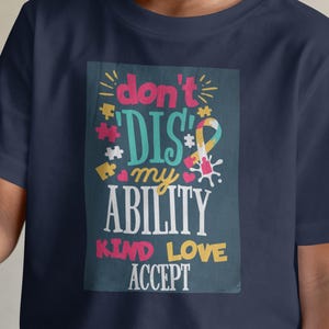 May include: Navy blue t-shirt with the text "don't DIS my ABILITY KIND LOVE ACCEPT" in a colorful design. The shirt features puzzle pieces and a ribbon. A child is smiling and holding a bouquet of flowers.