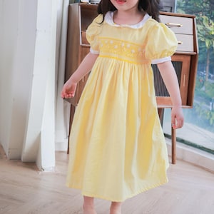 May include: A young girl wearing a yellow dress with a white collar and puff sleeves. The dress features a detailed bodice with floral embroidery. The girl is standing on a wooden floor in front of a window.