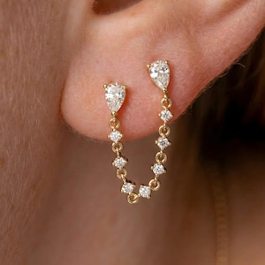 May include: A gold chain earring featuring two pear-shaped diamonds at the top, connected by a delicate gold chain adorned with six small round brilliant-cut diamonds, creating a draped effect between two piercing points on the earlobe.
