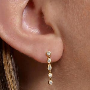 May include: A gold earring with five clear, round gemstones suspended in a vertical line. The earring is attached to the earlobe. The gemstones are evenly spaced and appear to be set in a gold metal.