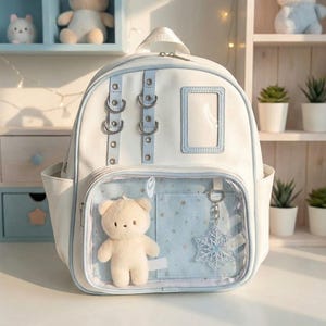 Op de afbeelding: Witte rugzak met lichtblauwe accenten, met een doorzichtige voorzak met een teddybeerknuffel en een sneeuwvlokbedel. De rugzak heeft zilverkleurige metalen hardware.