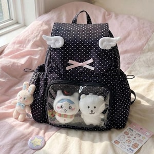 May include: Black polka dot backpack with white wing accents, a pink bow, and a clear front pocket displaying two plush toys. A plush bunny, a star pin, and a sticker sheet are also visible.