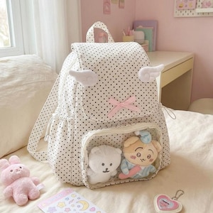 K&ouml;nnte beinhalten: Wei&szlig;er Rucksack mit Polka-Dots, Engelsfl&uuml;gel-Akzenten und einer rosa Schleife. Der Rucksack hat eine transparente Tasche mit zwei Pl&uuml;schtieren. Ein rosa Hasenpl&uuml;sch, ein Aufkleberbogen und ein herzf&ouml;rmiger Schl&uuml;sselanh&auml;nger sind ebenfalls sichtbar.