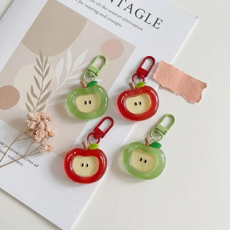 May include: Four apple-shaped keychains in red and green with a light-colored core. Each keychain has a metal clasp and a small green leaf. The keychains are on a white surface with a magazine and dried flowers.