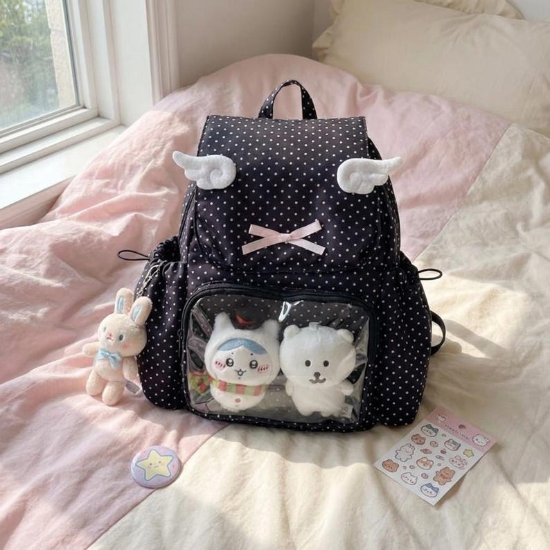 May include: Black polka dot backpack with white wing accents, a pink bow, and a clear front pocket displaying two plush toys. A plush bunny, a star pin, and a sticker sheet are also visible.