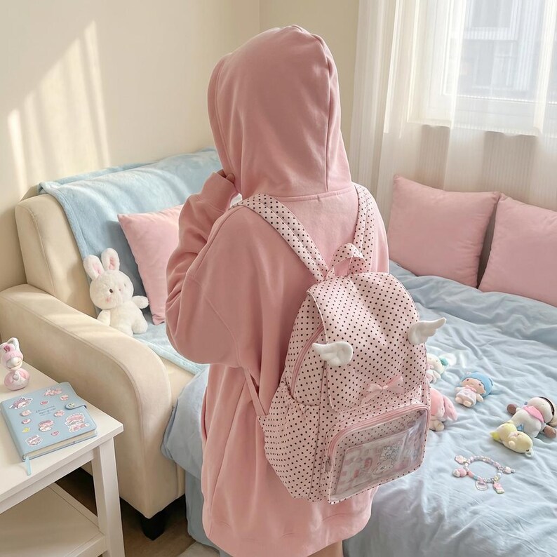 K&ouml;nnte beinhalten: Ein rosa Kapuzenpullover und ein gepunkteter Rucksack mit wei&szlig;en Fl&uuml;geln. Der Rucksack hat eine transparente Tasche und eine Schleife. Die Person befindet sich in einem Schlafzimmer mit einer hellblauen Tagesdecke und rosa Kissen.