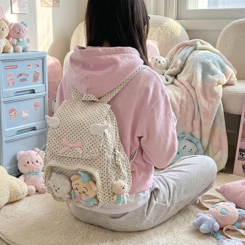 K&ouml;nnte beinhalten: Ein cremefarbener Rucksack mit Polka-Dots, Engelsfl&uuml;geln und einer rosa Schleife. Der Rucksack hat eine Vordertasche mit Pl&uuml;schtierfiguren. Die Person tr&auml;gt einen rosa Kapuzenpullover und eine graue Jogginghose.