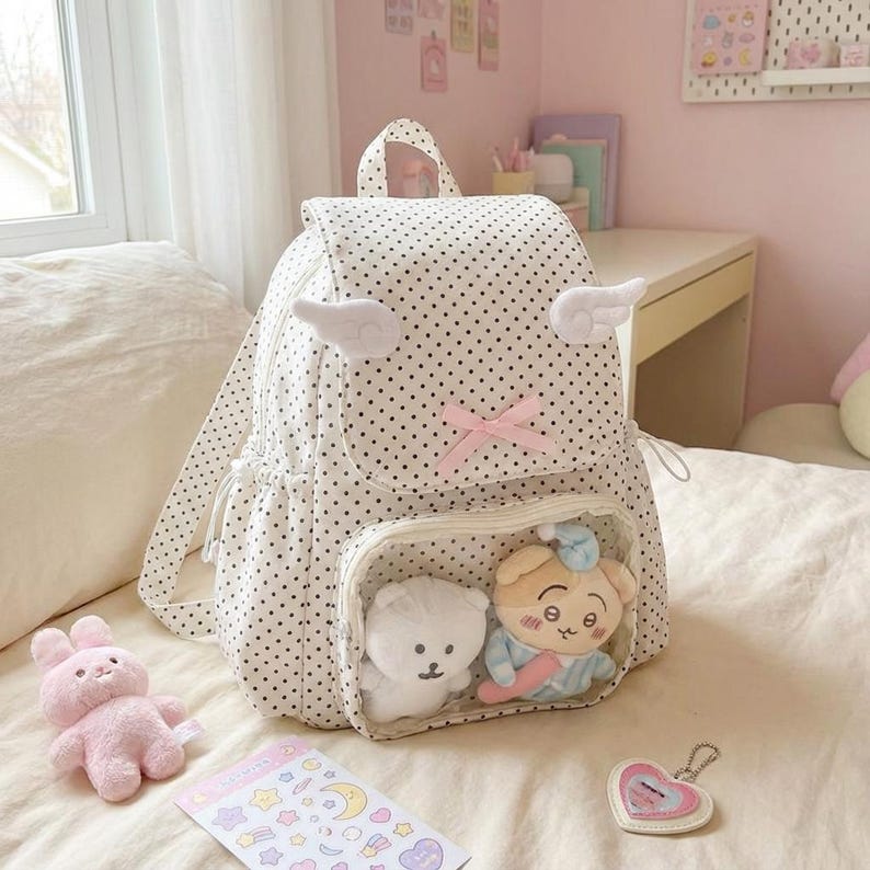 K&ouml;nnte beinhalten: Wei&szlig;er Rucksack mit Polka-Dots, Engelsfl&uuml;gel-Akzenten und einer rosa Schleife. Der Rucksack hat eine transparente Tasche mit zwei Pl&uuml;schtieren. Ein rosa Hasenpl&uuml;sch, ein Aufkleberbogen und ein herzf&ouml;rmiger Schl&uuml;sselanh&auml;nger sind ebenfalls sichtbar.