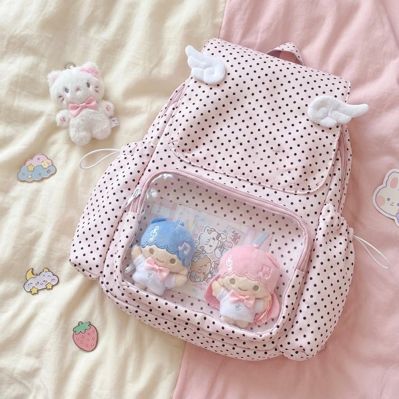 K&ouml;nnte beinhalten: Rosa Rucksack mit schwarzen Punkten, mit einer transparenten Vordertasche mit zwei Pl&uuml;schpuppen. Der Rucksack hat wei&szlig;e Fl&uuml;gelakzente und einen kleinen wei&szlig;en Katzen-Schl&uuml;sselanh&auml;nger. Dekorative Aufkleber sind verstreut.