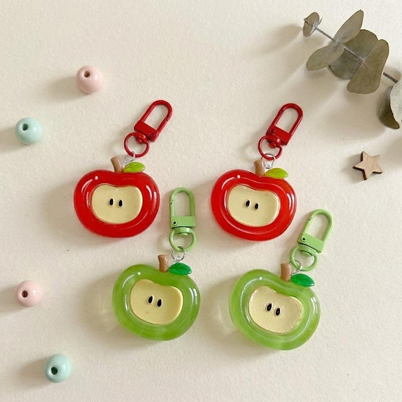 May include: Four apple-shaped keychains in red and green. The keychains have a white center with two small black dots, resembling apple seeds. Each keychain has a metal clasp and a small stem and leaf.