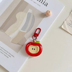 May include: A red apple-shaped keychain with a red clasp. The keychain has a green leaf and a tan-colored center with two small black dots. The keychain is on a white surface with a magazine.