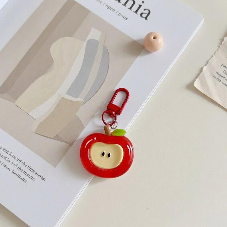 May include: A red apple-shaped keychain with a red clasp. The keychain has a green leaf and a tan-colored center with two small black dots. The keychain is on a white surface with a magazine.