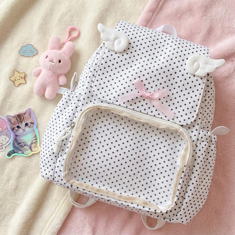 K&ouml;nnte beinhalten: Wei&szlig;er Rucksack mit schwarzen Punkten, einer transparenten Vordertasche und einer rosa Schleife. Der Rucksack hat wei&szlig;e Engelsfl&uuml;gel und einen rosa Hasen-Schl&uuml;sselanh&auml;nger. Ein Katzenaufkleber und andere kleine Anh&auml;nger sind ebenfalls sichtbar.