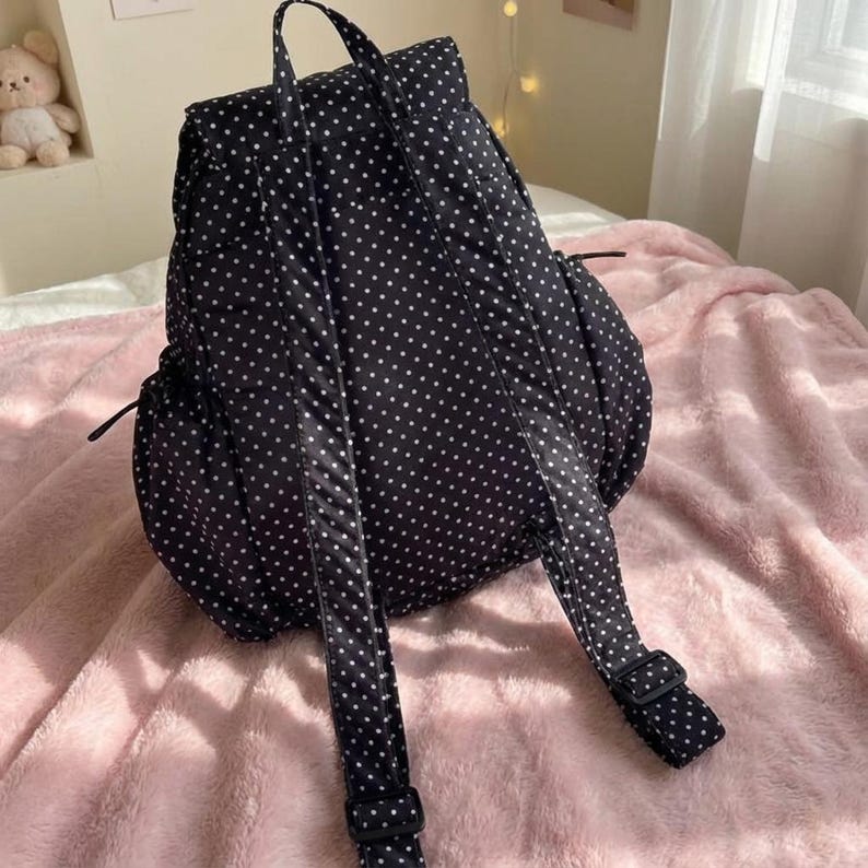 K&ouml;nnte beinhalten: Schwarzer Rucksack mit wei&szlig;en Polka-Dots. Der Rucksack hat verstellbare Tr&auml;ger und einen oberen Griff. Er hat eine Seitentasche und eine Frontklappe. Die Tasche liegt auf einer rosa Decke.