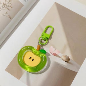May include: A green apple-shaped keychain with a yellow center and black seeds. The keychain has a green clasp and a small orange stem with a green leaf. The apple is made of a translucent material.