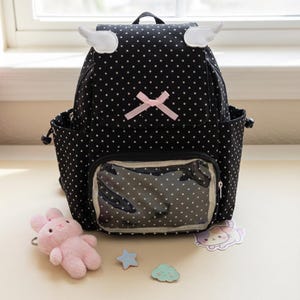K&ouml;nnte beinhalten: Schwarzer Rucksack mit wei&szlig;en Punkten, mit wei&szlig;en Fl&uuml;gelakzenten und einer rosa Schleife. Der Rucksack hat eine transparente Vordertasche und Seitentaschen. Ein rosa Pl&uuml;schhase, ein blauer Stern und ein Wolkenaufkleber liegen neben dem Rucksack.