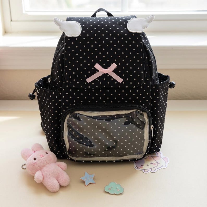 K&ouml;nnte beinhalten: Schwarzer Rucksack mit wei&szlig;en Punkten, mit wei&szlig;en Fl&uuml;gelakzenten und einer rosa Schleife. Der Rucksack hat eine transparente Vordertasche und Seitentaschen. Ein rosa Pl&uuml;schhase, ein blauer Stern und ein Wolkenaufkleber liegen neben dem Rucksack.