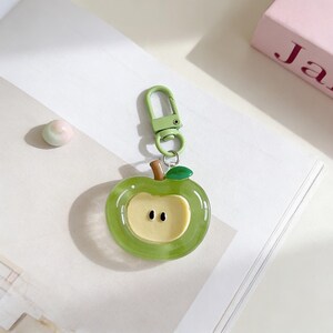 May include: A green apple-shaped keychain with a light green clasp and a brown stem. The apple has a light yellow center with two black dots. The keychain is on a white surface.