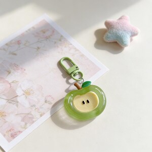 May include: A green apple-shaped keychain with a light green clasp and a brown stem. The apple has a yellow center with two black seeds. A small, fluffy star in pastel colors is in the background.