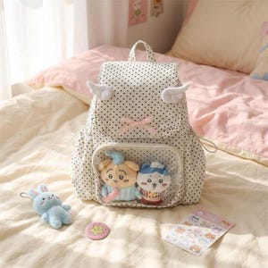 K&ouml;nnte beinhalten: Ein wei&szlig;er Rucksack mit schwarzen Punkten, Engelsfl&uuml;geln und einer rosa Schleife. Der Rucksack hat eine transparente Tasche mit zwei Pl&uuml;schtieren. Ein blaues Pl&uuml;schkaninchen, eine rosa sternf&ouml;rmige Anstecknadel und ein Aufkleberbogen sind ebenfalls sichtbar.