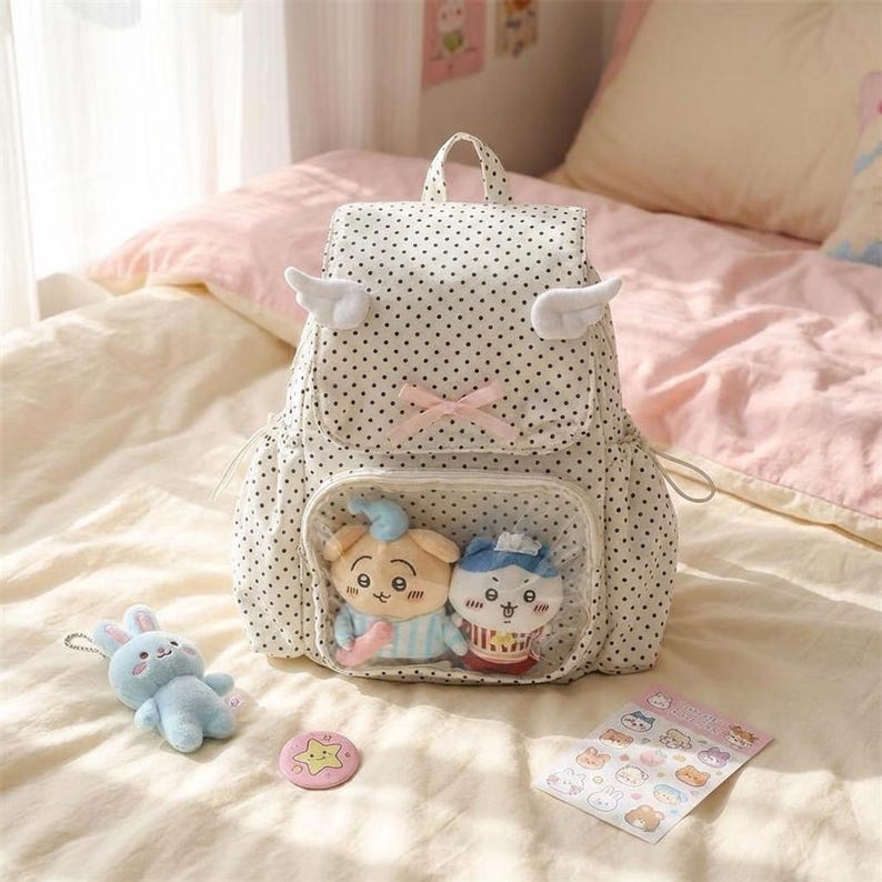 K&ouml;nnte beinhalten: Ein wei&szlig;er Rucksack mit schwarzen Punkten, Engelsfl&uuml;geln und einer rosa Schleife. Der Rucksack hat eine transparente Tasche mit zwei Pl&uuml;schtieren. Ein blaues Pl&uuml;schkaninchen, eine rosa sternf&ouml;rmige Anstecknadel und ein Aufkleberbogen sind ebenfalls sichtbar.