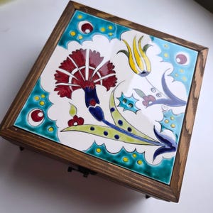 May include: Square wooden box with a decorative ceramic tile top. The tile features a floral design with a red flower, yellow tulip, and blue and green accents. The box has a dark brown wooden frame and a metal clasp.