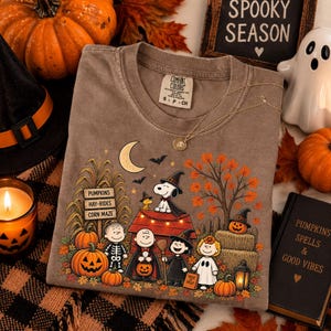 May include: A brown short-sleeved t-shirt featuring a graphic print of Peanuts characters in Halloween costumes. The design includes Snoopy on a doghouse, pumpkins, a crescent moon, bats, and a sign reading Pumpkins, Hay-Rides, Corn Maze.