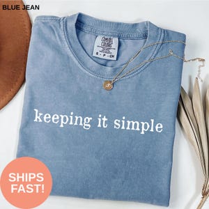 Könnte beinhalten: Ein blaues Jeans-T-Shirt mit dem weißen Schriftzug "keeping it simple". Eine goldene Halskette mit Anhänger liegt auf dem Shirt. Das Shirt hat ein Comfort Colors-Etikett.