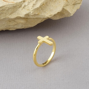 May include: A polished gold-tone ring featuring a small, centred cross emblem on the band. The ring has a smooth, reflective surface and a simple, circular design. The cross is positioned horizontally across the top of the thin, rounded band.
