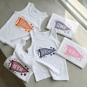 May include: Five white sleeveless tank tops featuring embroidered pennant designs. Each pennant displays a three-letter acronym: OAH in orange gingham, UMB in black gingham, UAH in navy gingham, VNU in pink gingham, and NYU in maroon gingham, with small icons.