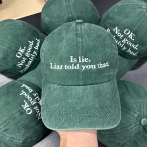 May include: A collection of green baseball caps with white embroidered text. One cap reads "Is lie. Liar told you that." Other caps say "OK. Not good. Probably bad." The caps are made of a soft, textured fabric.