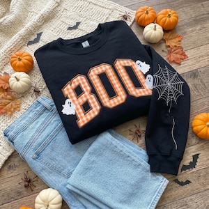 May include: A black long-sleeve sweatshirt featuring the word BOO in orange and white gingham letters. The design includes small white ghost graphics and a white spiderweb embroidery on the sleeve. The sweatshirt is paired with light blue denim jeans.