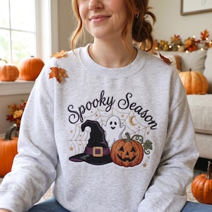 May include: A light gray crewneck sweatshirt featuring an embroidered design with the text Spooky Season, a black witch hat with a purple band, a white ghost, a carved orange pumpkin, a spider web, a crescent moon, and small gold stars.