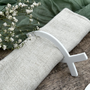 May include: A folded linen napkin is held by a white napkin ring. The napkin is a light beige color, and the napkin ring has a cross-like design. White flowers and green fabric are in the background.