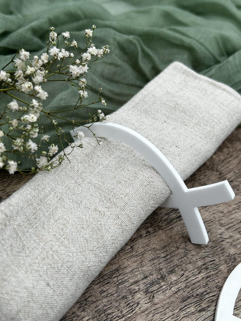 May include: A folded linen napkin is held by a white napkin ring. The napkin is a light beige color, and the napkin ring has a cross-like design. White flowers and green fabric are in the background.