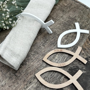May include: A white fish-shaped napkin ring holds a beige linen napkin on a wooden tray. Two wooden fish-shaped place cards are visible, one with the name "MAGDALENA" and the date "21.08.2024" engraved.