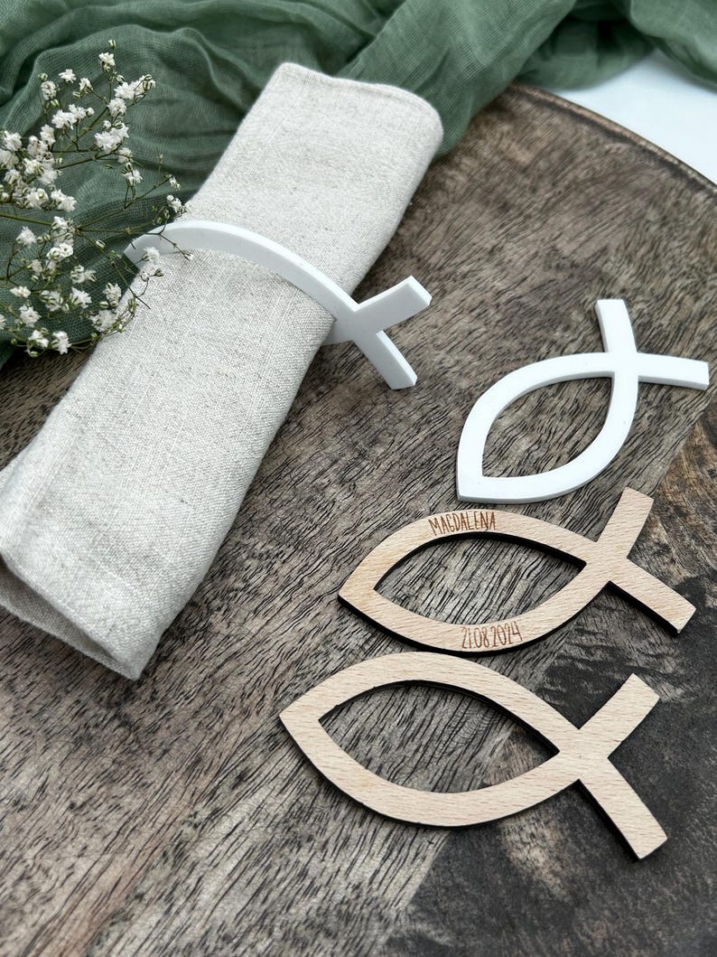 May include: A white fish-shaped napkin ring holds a beige linen napkin on a wooden tray. Two wooden fish-shaped place cards are visible, one with the name "MAGDALENA" and the date "21.08.2024" engraved.