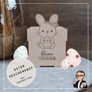 Op de afbeelding: Een houten paascadeaudoos met een lasergeëtste konijn met een ei. De doos heeft de tekst "kleiner OSTERGRUB" op de voorkant. Twee decoratieve paaseieren staan naast de doos. Een ronde sticker zegt "OSTER GESCHENKBOX".